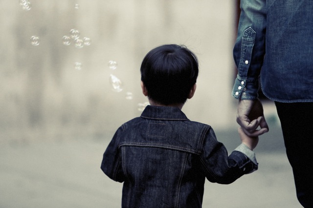 A young child in a denim jacket holds an adult's hand while watching soap bubbles float in the air, with a blurred background emphasizing a serene parenting time moment.