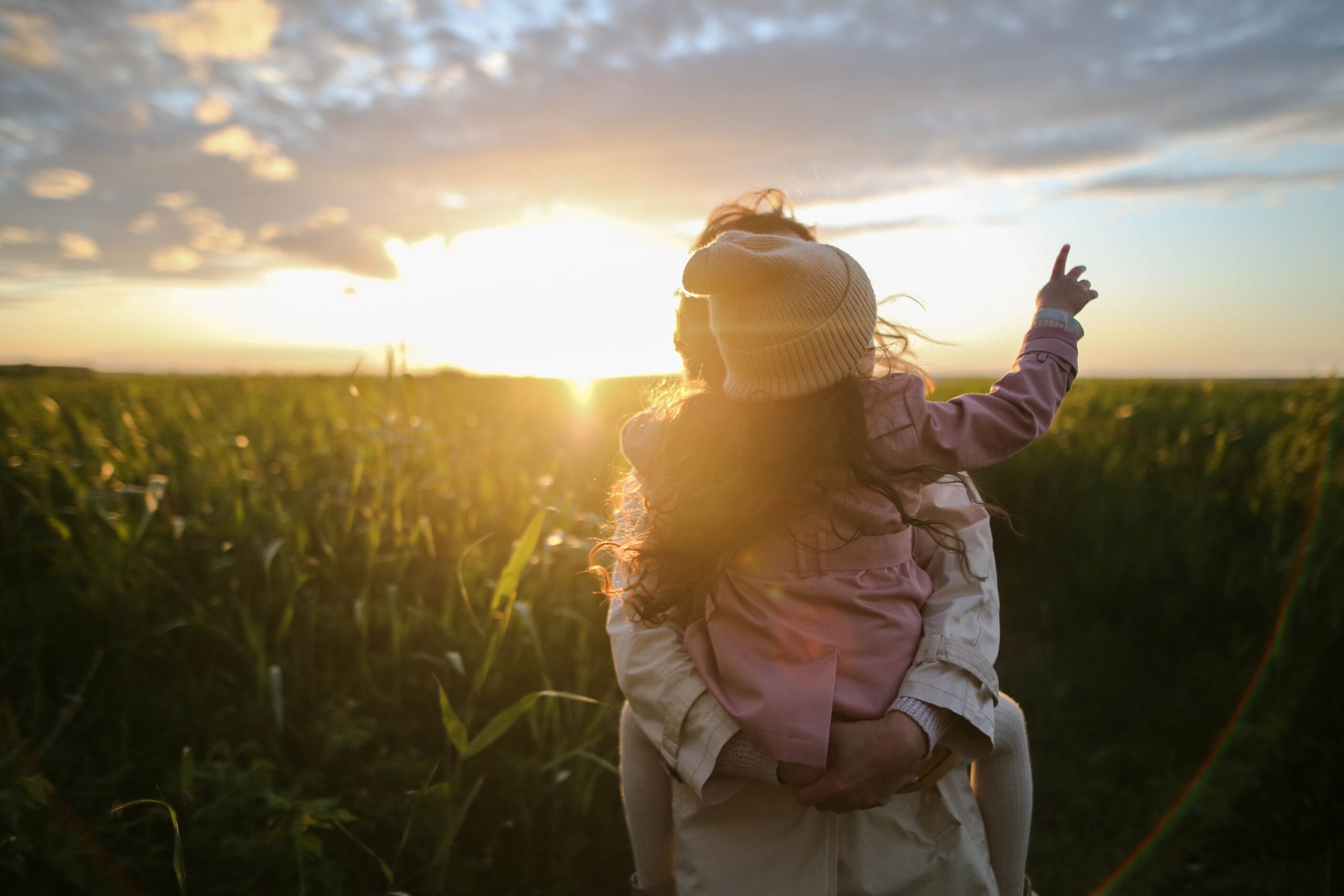 A young child in a beanie and jacket is being lifted up by an adult in a field in the State of New Jersey at sunset, pointing towards the sky as the sun casts a warm glow around