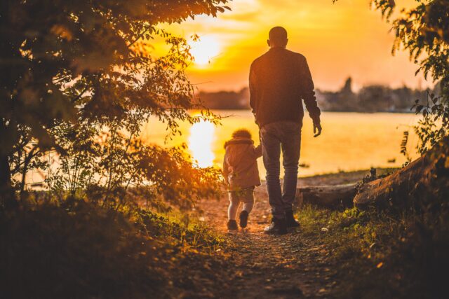 A man and a small child walk together beside a lake in New Jersey at sunset, surrounded by golden light and autumn foliage, creating a serene and heartwarming scene.