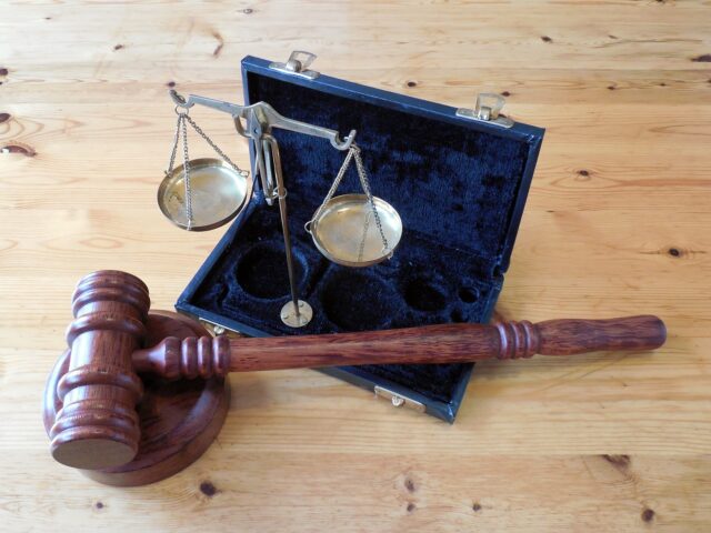 A wooden gavel, often used to affect divorce proceedings, lies next to an open small suitcase displaying a brass balance scale, set on a light wooden table.