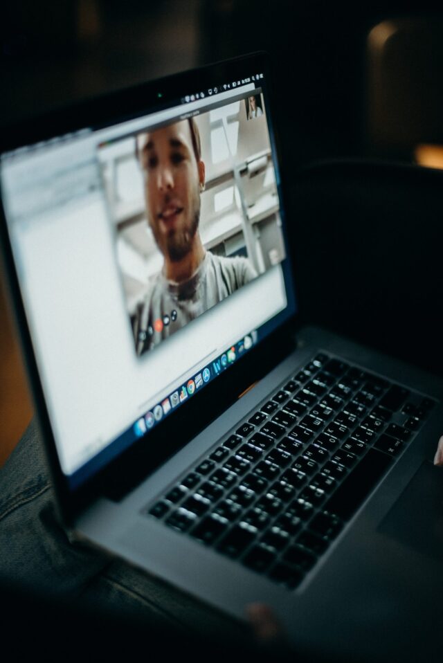 A close-up view of an open laptop showing a virtual visit with a smiling young man on the screen. The laptop is on a wooden surface, illuminated by soft light.