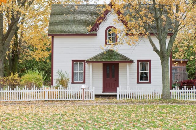 A quaint white house with a red door and autumn leaves scattered on the ground, surrounded by a white picket fence, with trees displaying fall colors in the background, to be kept in the NJ divorce