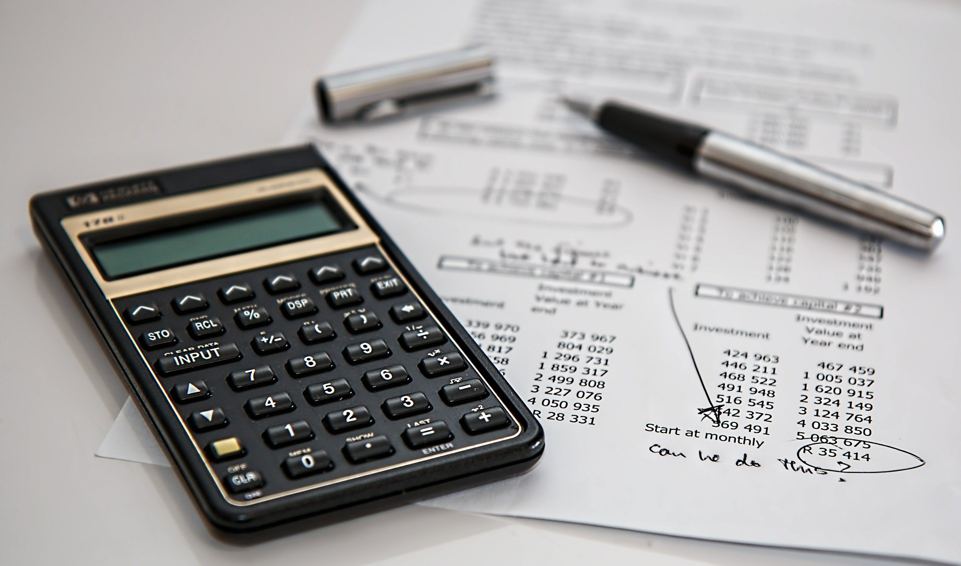 A calculator, pen, and several papers filled with numerical data and handwritten calculations are shown on a desk, highlighting the work of a forensic accountant.