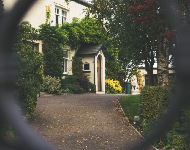 View through a circular frame of a charming house with ivy-covered walls, perfect for relocation, surrounded by lush gardens and a paved driveway. There's a statue visible near the vibrant foliage.