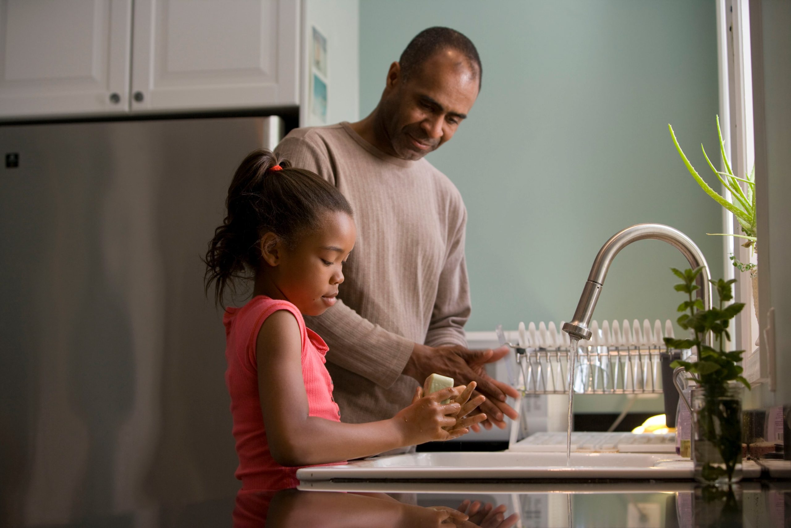 A father and his young daughter, who he shares in child custody, wash dishes together in a kitchen. He smiles watching her as she scrubs a plate carefully by the sink.