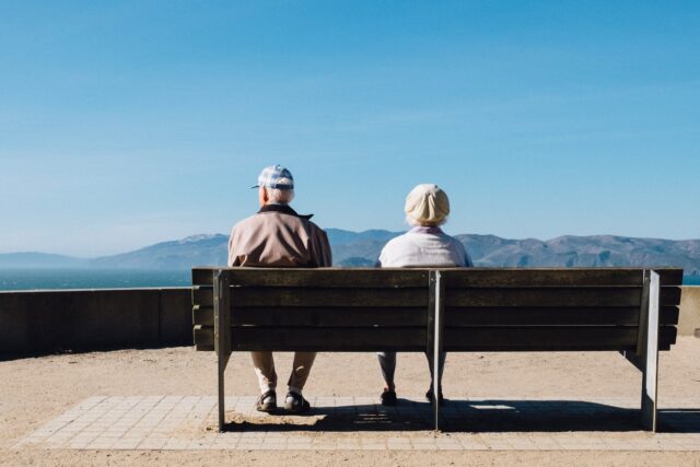 Two elderly people, amidst a gray divorce, sitting on a bench overlooking a scenic mountain range under a clear blue sky.