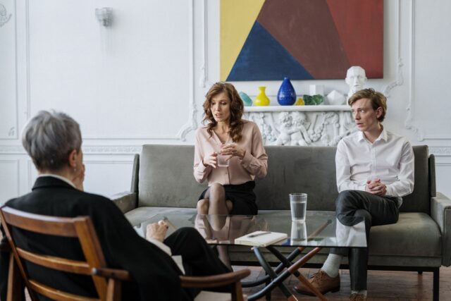 Two people sitting on a couch in an elegantly decorated room with a third person seated across from them in a chair, listening intently during a mediated divorce discussion.