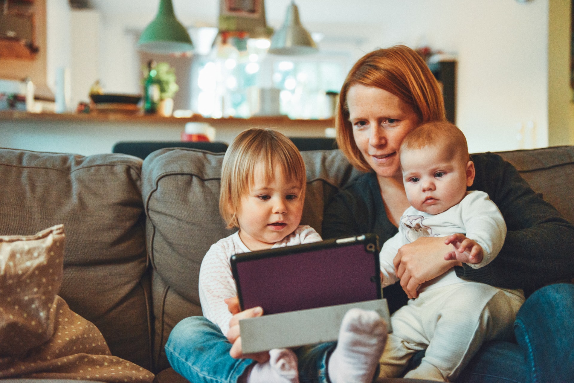 A mother sits on a couch with her two young children, showing a tablet to the older child while holding the younger one. They seem engaged and content in a cozy living room setting during the holidays.