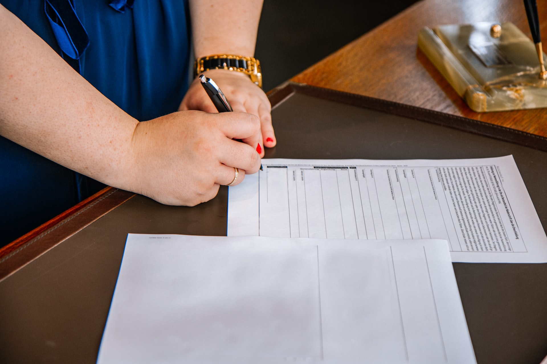A person with red nail polish is signing a prenuptial agreement at a wooden table, with more papers and a pen holder visible.