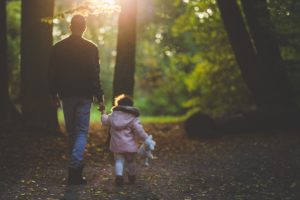 A man and a young child walk hand-in-hand through a forest, bathed in soft sunlight filtering through the trees. The child carries a toy, symbolizing the cherished moments and rights upheld in New