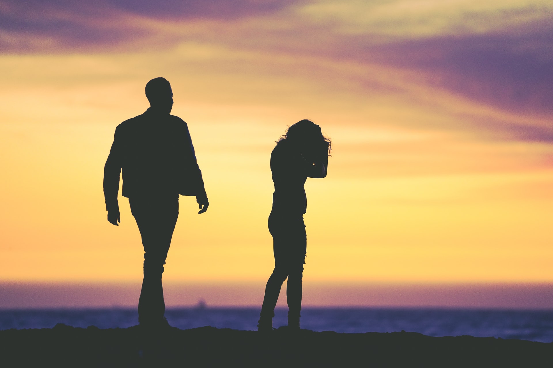 Silhouetted man and woman, both in their early 20s, stand apart on a beach at sunset, with a vibrant purple and orange sky in the background.